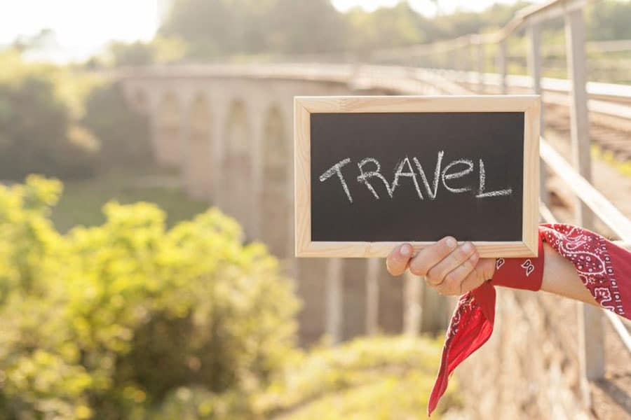 A sign held by a hand that says Travel written with chalk on the board.