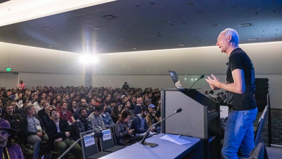 a male person giving speech about GDC 2025 on a hall full of people.